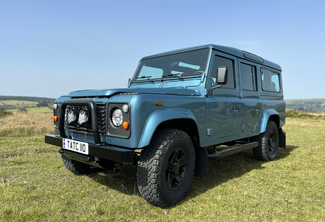 A Land Rover Defender 110 is parked in a layby area in the countryside with a scenic hill in the background. 