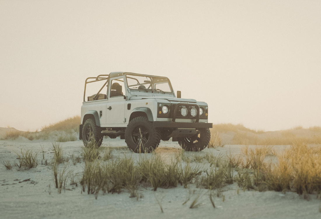 A white Land Rover Defender sits on a sunny beach with its tire marks showing in the sand. 