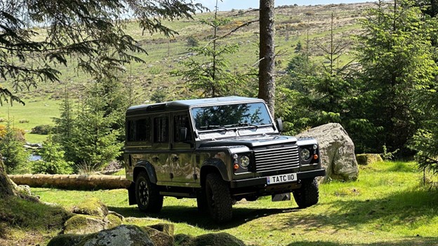 A Land Rover Defender 110 sits on a grass path on the edge of a forest, it is overlooked by a big hill.