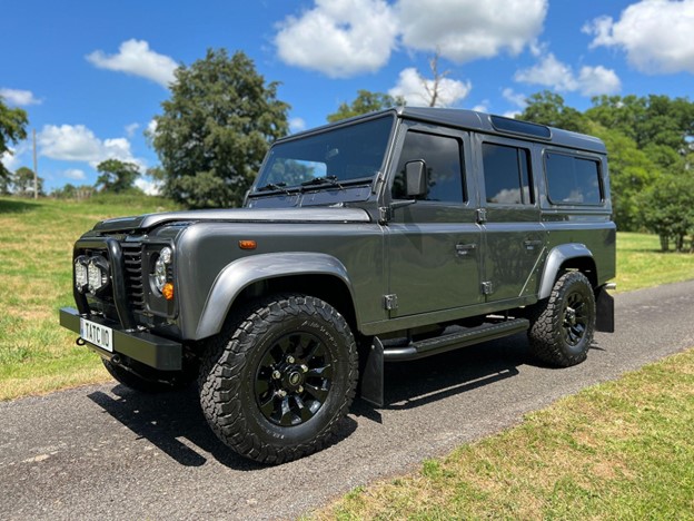 A Land Rover Defender 110 is parked on a through road in the grounds of a country park.